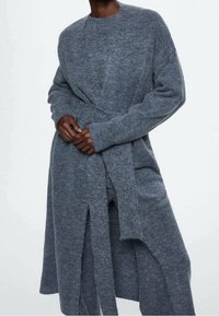 Długi, szary kardigan z dzianiny z okrągłym dekoltem, długimi rękawami i paskiem do wiązania w talii. Charakteryzuje się luźnym krojem i bocznymi rozcięciami.