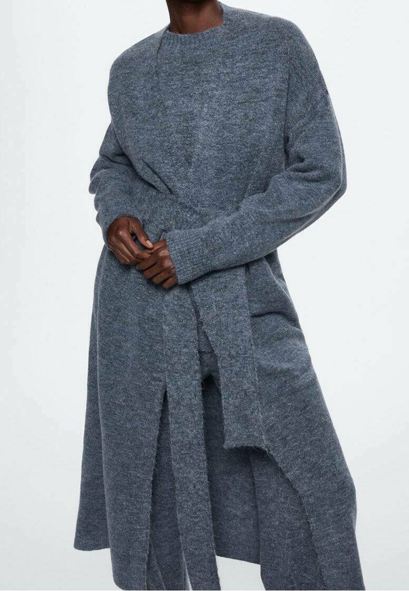 Długi, szary kardigan z dzianiny z okrągłym dekoltem, długimi rękawami i paskiem do wiązania w talii. Charakteryzuje się luźnym krojem i bocznymi rozcięciami.