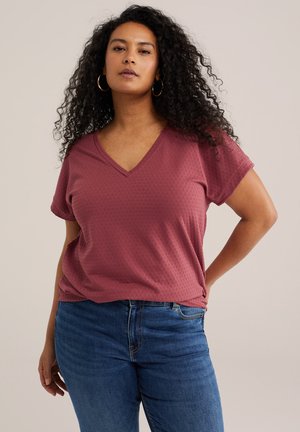 Žena s dlhými kučeravými vlasmi, ktorá má náušnice kruhy, ružový top s krátkym rukávom a výstrihom do V a modré rifle, stojí s jednou rukou na boku.