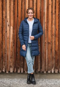 Cappotto imbottito blu navy con cappuccio, design a righe verticali, chiusura con zip, indossato sopra un maglione grigio chiaro con motivo a treccia, abbinato a jeans con risvolto e stivaletti neri.