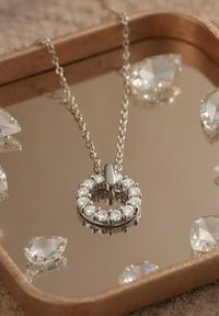 Silverhalsband med en cirkulär hänge prytt med flera klara stenar, som vilar på en reflekterande yta med spridda diamantformade detaljer.