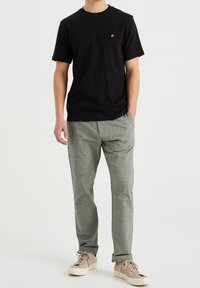 Czarna bawełniana koszulka z złotym logo na klatce piersiowej, w połączeniu z szarymi, teksturowanymi spodniami i beżowymi trampkami. Luźny krój i podwinięte mankiety.