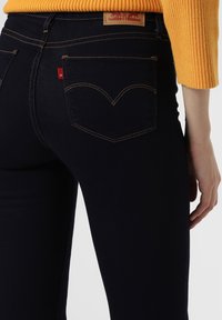 Jeans de mezclilla azul oscuro con un diseño ajustado, que presentan costuras amarillas en contraste y una etiqueta de cuero con un logo en la parte trasera de la cinturilla.