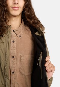 Persoon met lang krullend haar, gekleed in een tan corduroy overhemd, houdt een olijfgroene jas open om de zwarte fleece-voering aan de binnenkant te tonen.