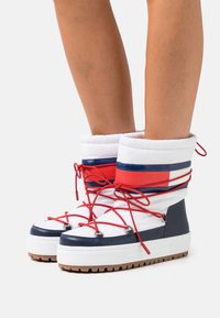 Białe i granatowe buty śniegowe z czerwonymi akcentami, wyposażone w wypchaną cholewkę, okrągły czubek i grubą gumową podeszwę. Sznurówki przechodzą przez metalowe oczka.