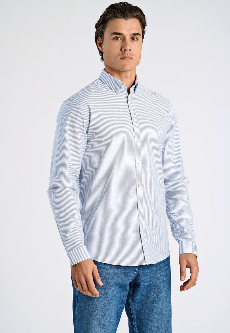 Mann trägt hellblau-weiß gestreiftes Hemd mit langen Ärmeln und blaue Jeans, steht vor einfachem Hintergrund.