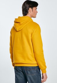Żółta hoodie z miękką teksturą, z prążkowanymi mankietami i dołem, noszona na ciemnych dżinsach. Posiada kieszeń kangurzą i luźny krój.