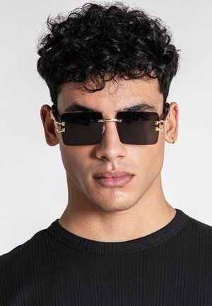 Mand med krøllet sort hår iført rektangulære sorte solbriller med guldaccents og en lille gylden hoop-ørering, iført en sort ribbet skjorte.