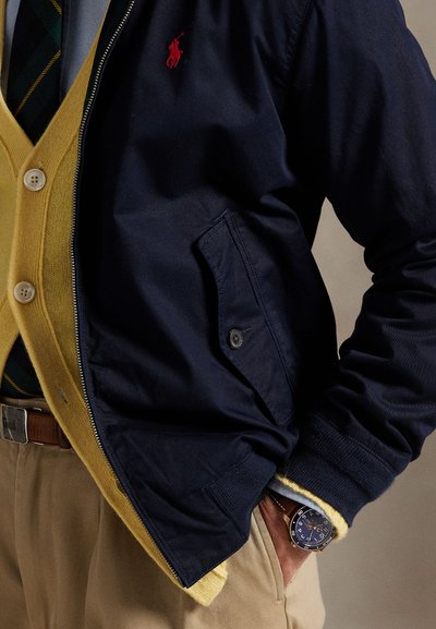 Bărbat purtând o jachetă bleumarin cu fermoar peste un cardigan galben, cravată cu dungi, pantaloni bej și un ceas de mână cu față bleumarin și curea din piele maro.