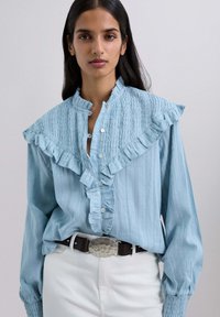 Blusa azul claro con cuello y tapeta con volantes, tela texturizada, cierre de botones y mangas largas abullonadas, llevada con un cinturón oscuro.