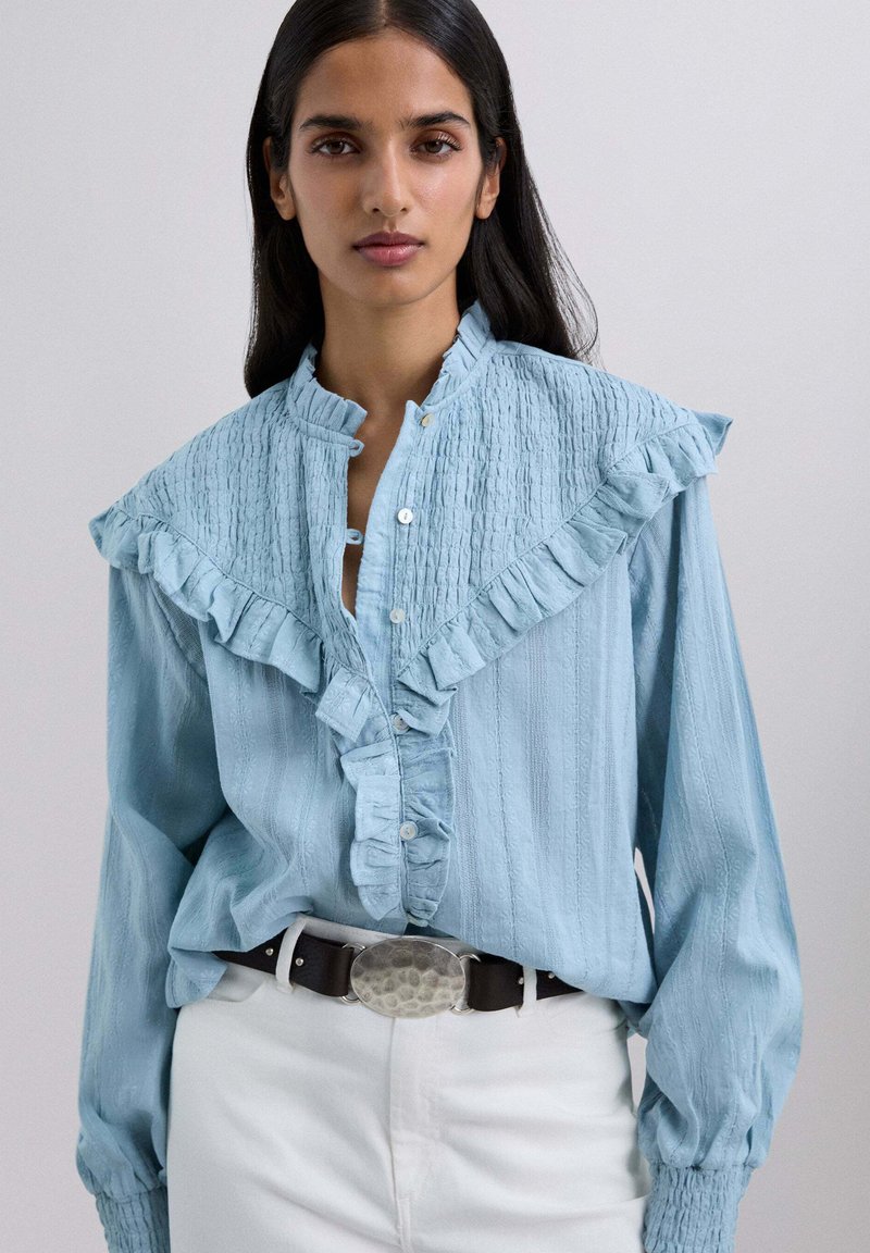 Blusa azul claro con cuello y tapeta con volantes, tela texturizada, cierre de botones y mangas largas abullonadas, llevada con un cinturón oscuro.