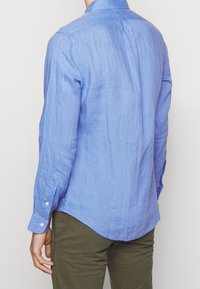Light blue linen shirt with long sleeves, curved hem, buttoned cuffs, and subtle texture. The back features a center pleat for shape.