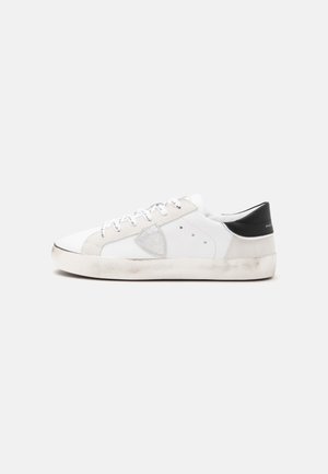 Baskets basses blanches avec empiècement noir au talon, lacets blancs, patch gris sur le côté et semelle blanc cassé légèrement sale sur fond uni.