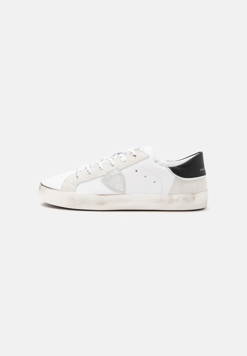 Baskets basses blanches avec empiècement noir au talon, lacets blancs, patch gris sur le côté et semelle blanc cassé légèrement sale sur fond uni.
