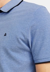 Lysblå polo-shirt lavet af tekstureret stof, med en sort detalje på kraven og et lille logo på brystet.