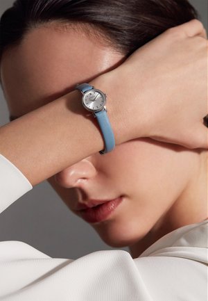 Montre-bracelet en argent avec un cadran blanc et un bracelet en cuir bleu, présentant une finition texturée et des accents en cristal sur la lunette.