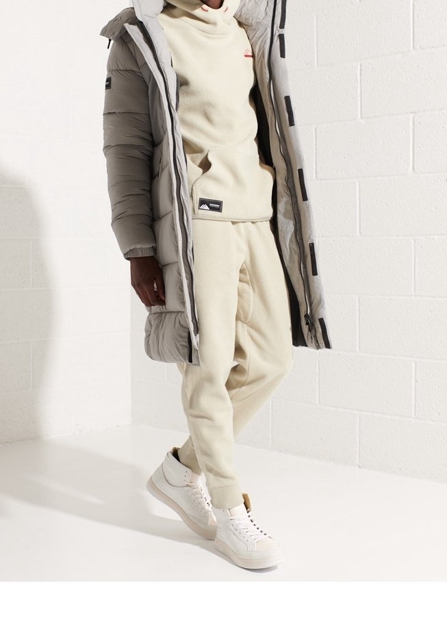 Personne portant un survêtement en polaire beige, un manteau matelassé gris et des baskets montantes blanches, debout contre un mur en briques blanches.