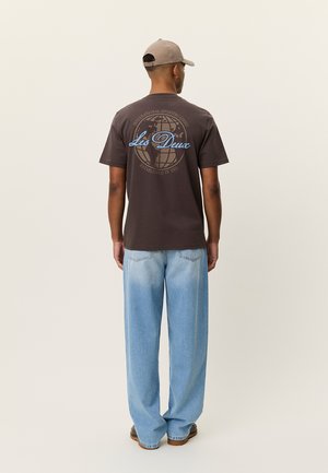 Braunes Baumwoll-T-Shirt mit einer Globus-Grafik und Text auf der Rückseite, kombiniert mit hellblauen Jeans mit weitem Bein und braunen Schuhen.
