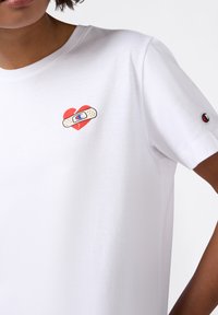 Persona che indossa una maglietta bianca con una grafica di un cuore spezzato rosso e una benda vicino al petto, e un piccolo logo sulla manica.
