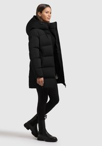 Abrigo acolchado negro con capucha, caracterizado por un diseño acolchado y puños elásticos. Llevado con pantalones negros y botas ankle.