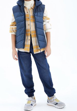 Polstret marineblå vest over en ternet creme- og gul skjorte, kombineret med løse blå cowboybukser og grå sneakers med gule detaljer.