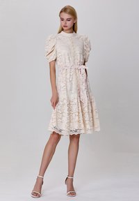 Robe en dentelle crème, avec des manches bouffantes, un bouton de fermeture et une ceinture à nouer à la taille. Fini avec un ourlet festonné.