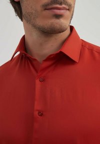 Chemise rouge à boutons avec une texture lisse, un col classique et deux boutons visibles au cou ; le tissu semble léger et élégant.