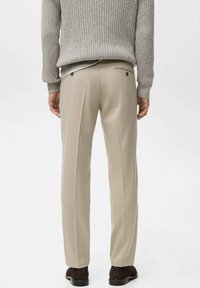 Pantalon beige sur mesure en tissu lisse, avec des plis doubles et des poches arrière, assorti à un pull gris texturé.