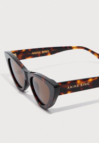 Kaķacs acs tumšu šoļu saulesbrilles ar bruņurupuču krāsas ietvaru un zelta "ANINE BING" logotipu uz briļļu kājiņām.