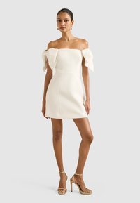 Bijela haljina bez ramena s velikim mašnama na rukavima, uska silueta, glatka tekstura i dužina iznad koljena. Zlate sandale s visokom petom.