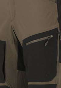 Close-up af todelt stofbukser med en lynlåslomme med trækflap og trekantet logo på den sorte sektion.