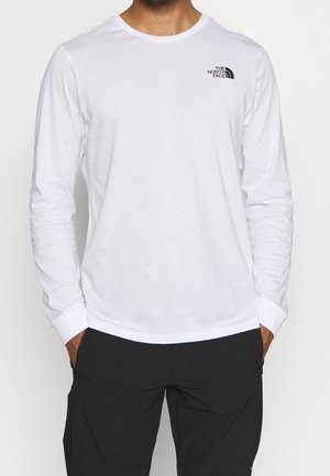 Homme portant un t-shirt blanc à manches longues The North Face et un pantalon noir, les mains dans les poches, debout devant un fond blanc.