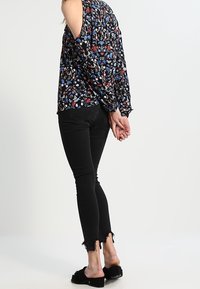 Blusa nera a fantasia floreale con design a spalle scoperte, abbinata a jeans skinny neri con orli sfrangiati e scarpe slip-on nere.