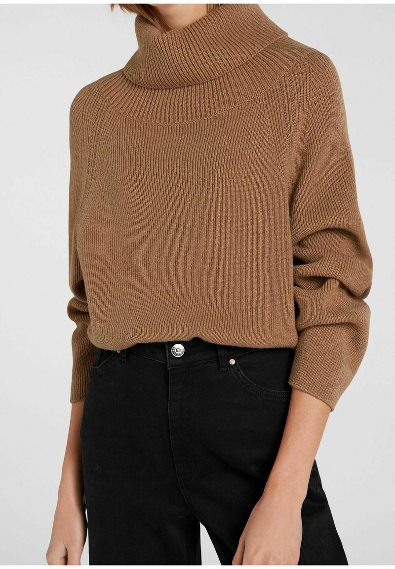 Pull à col roulé côtelé marron avec manches longues, associé à un jean noir taille haute portée par une personne sur un fond uni.