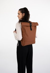Rucsac maro cu partea superioară rulată, fabricat dintr-un material neted, prezentând o cataramă și curele negre. Designul include buzunare laterale și o estetică minimalistă.