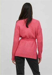Růžový pletený kardigan s dlouhými rukávy, detailem pásku v pase a žebrovanou texturou, kombinovaný s tmavými kalhotami.