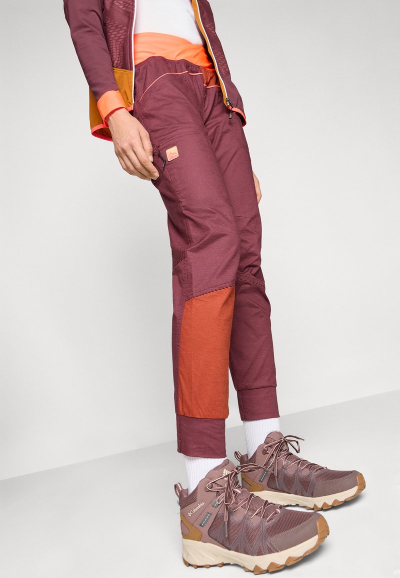 Bordeauxrote und orange Hosen mit Taschen und verstellbaren Bündchen. Braune Wanderschuhe mit Mesh- und Gummidetails. Weiße Socken sichtbar.