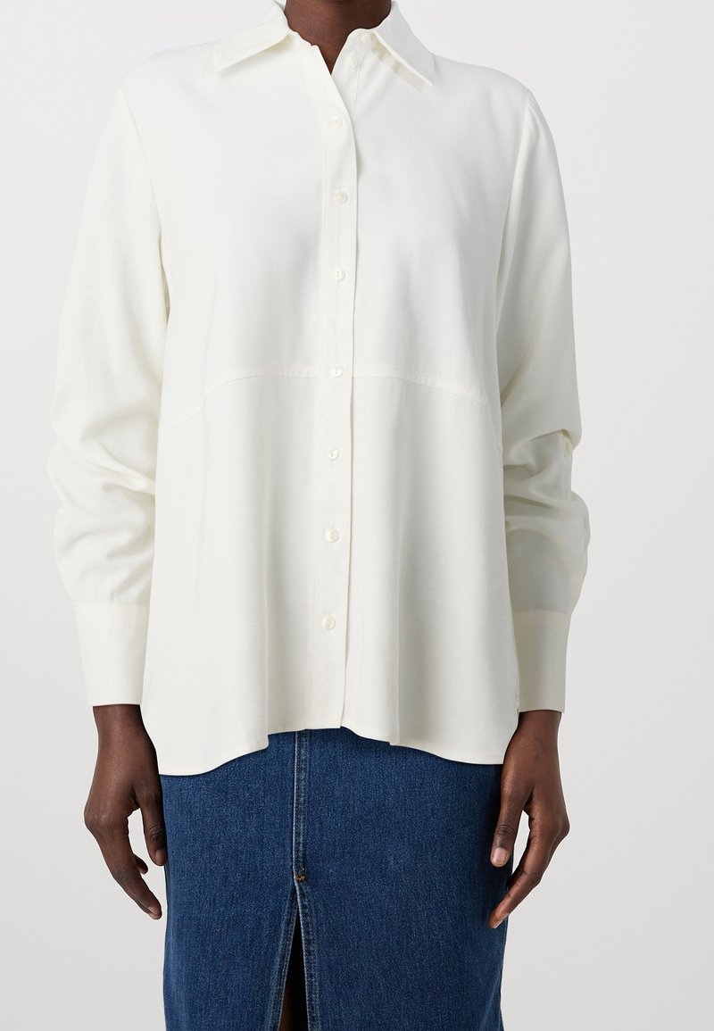 Chemise blanche à manches longues boutonnée, portée avec une jupe en jean bleu présentant une fente à l'avant, montrée des épaules jusqu'à mi-cuisse.