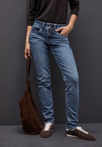 Jeans de color azul claro con un diseño ajustado, cinco bolsillos, un sutil desgastado y un ligero detalle rasgado, combinados con zapatos de ante marrón y un bolso.