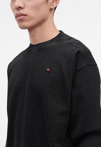 Jeune homme portant un sweat-shirt noir à col rond avec un petit logo rouge et blanc sur la poitrine gauche, sur un fond uni.