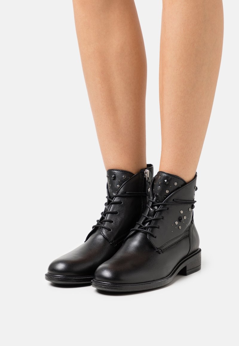 Bottines en cuir noir à lacets sur le devant avec des clous argentés décoratifs portées sur jambes nues.