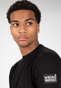 T-shirt in cotone nero con collo rotondo e logo ricamato "WRONG FRIENDS" sulla manica sinistra, caratterizzato da una texture liscia.