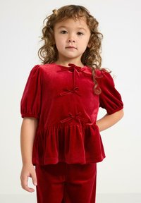 Jong meisje met krullend haar, gekleed in een rood fluwelen top met strikken en opbollende korte mouwen, poseert met één hand op de heup.