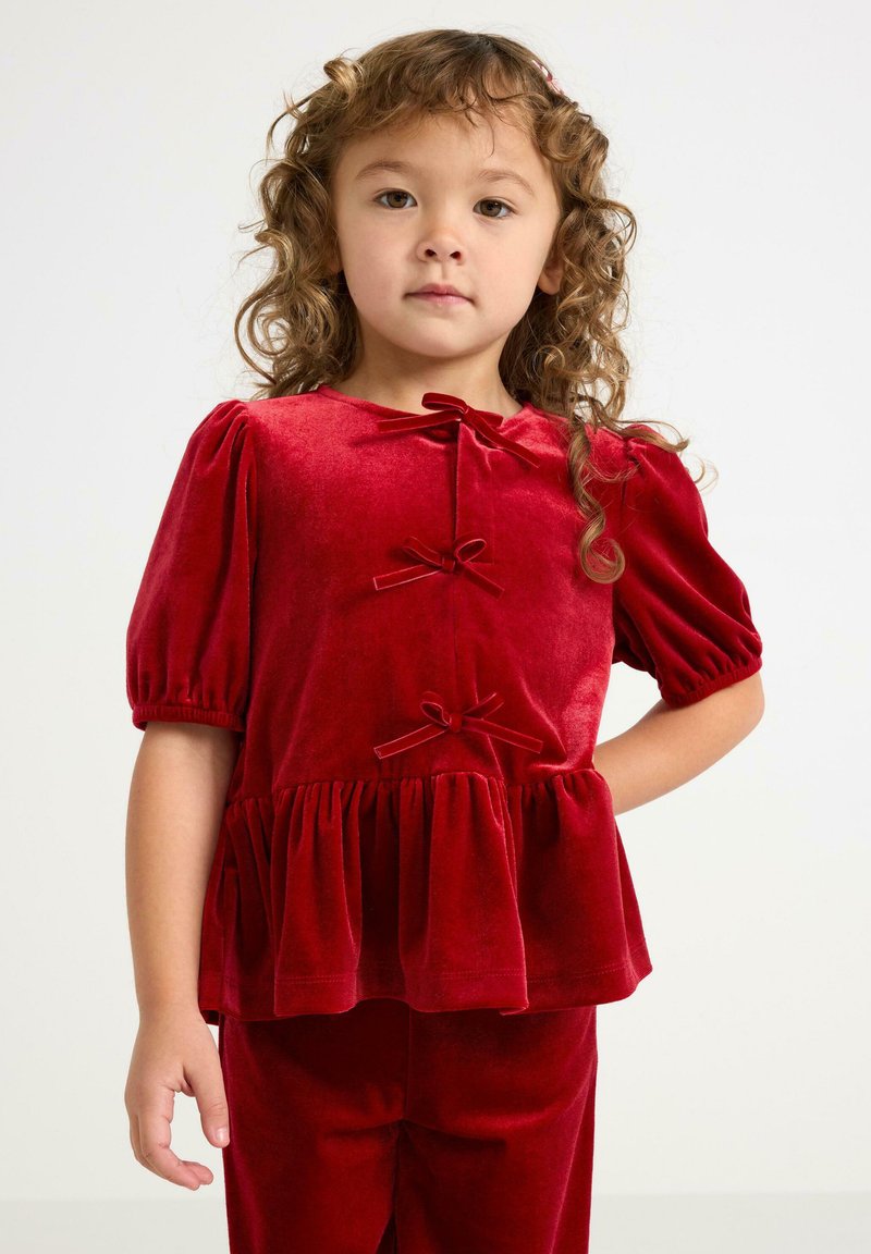 Jong meisje met krullend haar, gekleed in een rood fluwelen top met strikken en opbollende korte mouwen, poseert met één hand op de heup.