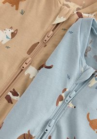 Deux grenouillères pour bébé avec fermeture éclair à l'avant, l'une beige et l'autre bleu clair, toutes deux ornées de diverses illustrations de petits chiens.