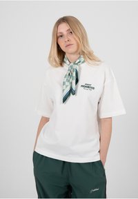 Hvit oversized t-skjorte med «PROHIBITED»-logo i svart, brukt med et mønstret skjerf i sjøgrønt og marineblått knyttet rundt halsen. Grønne treningsbukser.