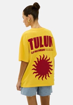 Camiseta amarilla de gran tamaño con un gran gráfico de sol rojo y el texto "TULUN", combinada con pantalones cortos de mezclilla azul claro.