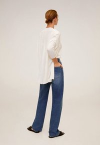 Bílý pletený top s jemným ribbed vzorem, s překrývající se zadní částí, kombinovaný s volnými modrými džíny. Modelka stojí uvolněně s rukama v kapsách. Černé ploché boty.