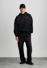 Μαύρη κουκούλα με κόκκινο λογότυπο, χαλαρή εφαρμογή, μπροστινή τσέπη; ταιριαστά μαύρα φόρμα με λογότυπο. Όλα τα είδη είναι κατασκευασμένα από μαλακό, υφασμάτινο υλικό.