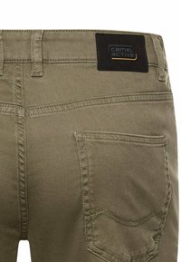 Rückansicht einer khakifarbenen Hose, die eine einzelne Tasche mit detaillierter Naht und einem schwarzen Stoffetikett mit dem Text "camel active" über der Gürtelschlaufe zeigt.